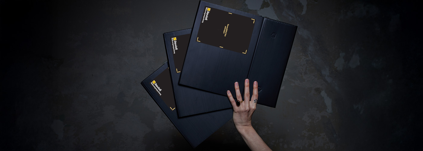 ravioli-reusable-shipping-boxes-folded 2.jpg A hand holding three collapsed, flat-folded black Ravioli reusable shipping boxes against a dark textured background, highlighting efficient return logistics